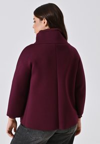 Vrouw met lang bruin haar, gekleed in een donkerpaarse jas met hoge kraag en lange mouwen, en donkergrijze jeans, met haar rug naar de camera toegekeerd.