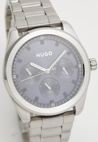 Montre en acier inoxydable argenté avec un cadran gris, trois sous-cadrans pour la date et le jour, et de grands marqueurs d'heure ; elle est dotée d'un bracelet en métal brossé.