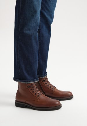 Brązowe skórzane botki za kostkę o gładkiej fakturze, z zaokrąglonym noskiem i czarnymi podeszwami. W połączeniu z ciemnoniebieskimi dżinsami, lekko podwiniętymi na mankietach.