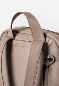 Tasche aus braunem Leder mit strukturierter Oberfläche, ausgestattet mit einem runden Griff und einem sichtbaren Reißverschluss. Ein Anhänger zeigt den Markennamen, dezent eingeprägt.