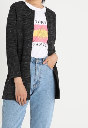 Kobieta ubrana w ciemnoszary kardigan z otwartym przodem na biały t-shirt z grafiką Tokio oraz jasnoniebieskie, wysokowysokie jeansy, z ręką w kieszeni.