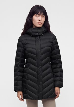 Femme portant un manteau matelassé noir avec un col montant et des boutons-pression, debout devant un fond clair uni.