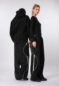 Fekete kapucnis pulóver és fekete, széles szárú nadrág fehér ívelt díszítésekkel és csíkokkal. Sima textúra, laza városi esztétika.