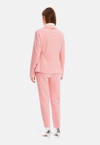 Rosa Anzug aus strukturiertem Stoff, bestehend aus einem taillierten Blazer und schmal geschnittenen Hosen. Die Rückansicht zeigt ein einzelnes Knopfdetail und einen weißen Kragen.