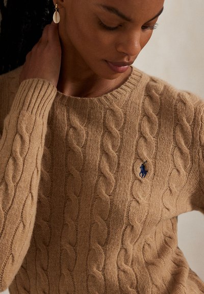 Femme portant un pull beige à torsades avec un logo de cheval brodé en bleu marine, touchant son cou d'une main, la tête inclinée vers le bas.
