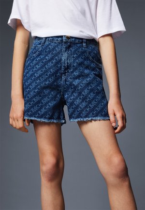 Persoon draagt hoog getailleerde blauwe denimshorts met een subtiel all-over logopatroon en rafelige zomen, gecombineerd met een eenvoudig wit T-shirt.