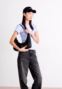 Bolso negro de hombro con un diseño curvado, textura suave y correa ajustable, combinado con una camisa ajustada de color azul claro y jeans negros.