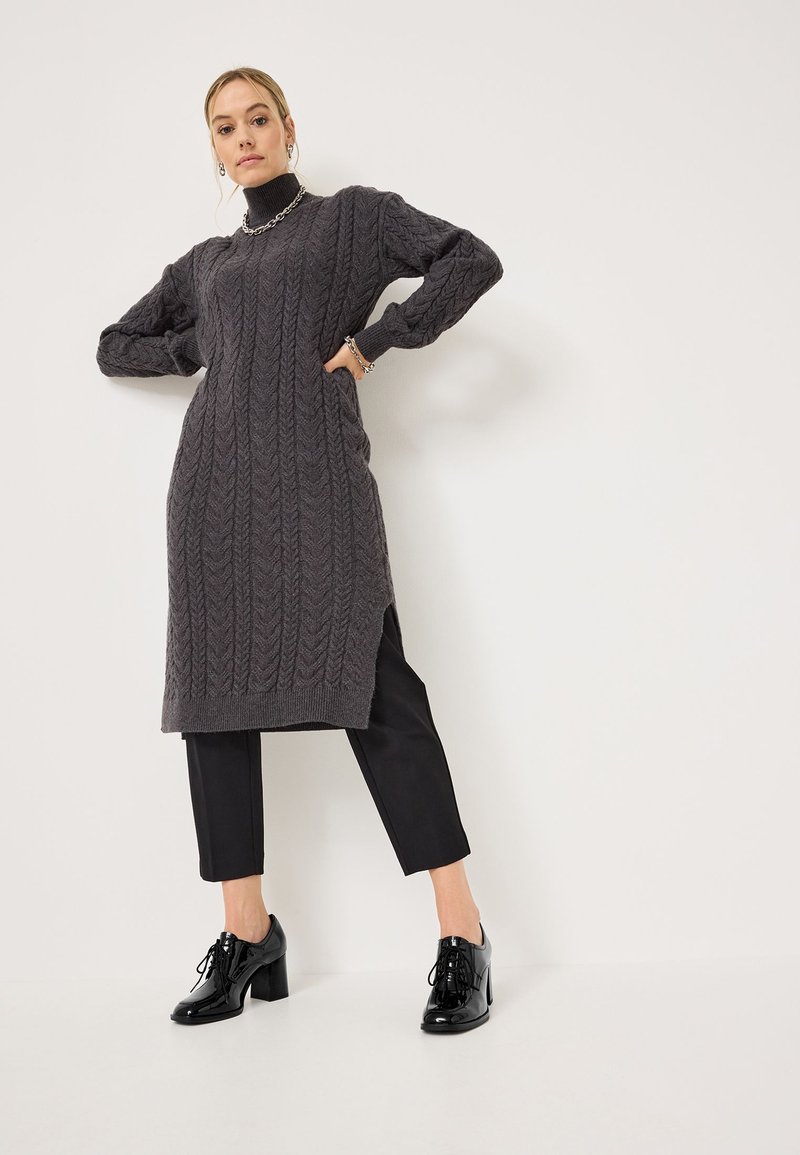 Abito maglione grigio scuro con motivo a treccia, maniche lunghe, spacchi laterali, abbinato a pantaloni cropped neri e scarpe con tacco lucide.
