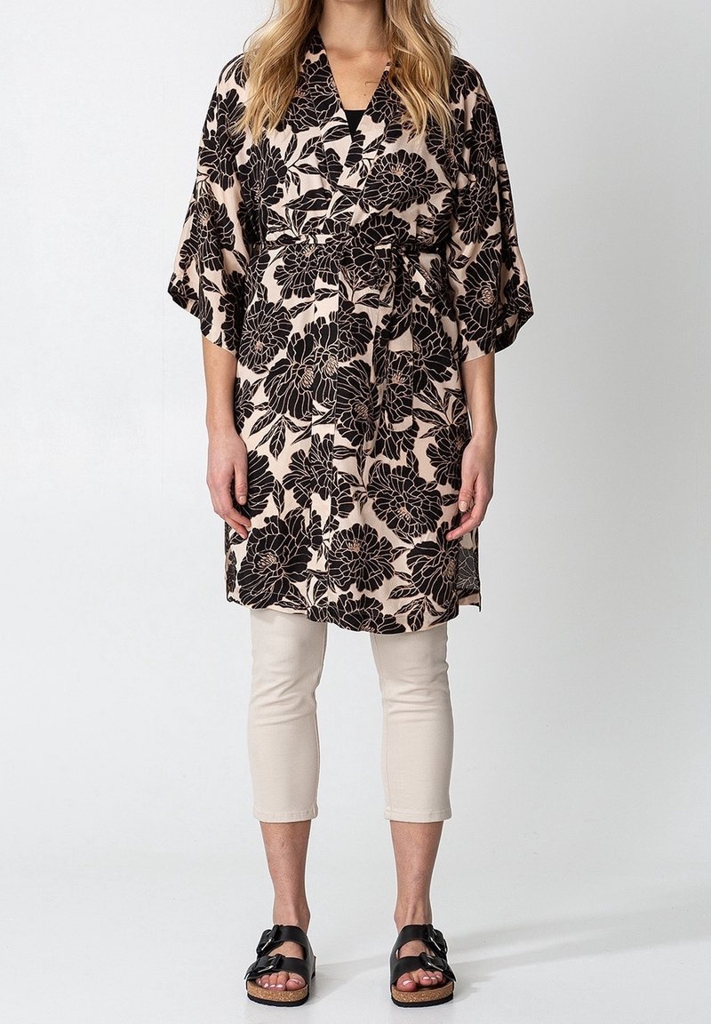 Femme portant un pantalon beige court, des sandales noires et une robe portefeuille beige à fleurs noires avec des manches larges.