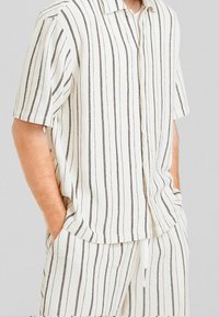 Man som bär en vit kortärmad skjorta med knappar och matchande shorts med tunna vertikala svarta ränder, med händerna i fickorna.