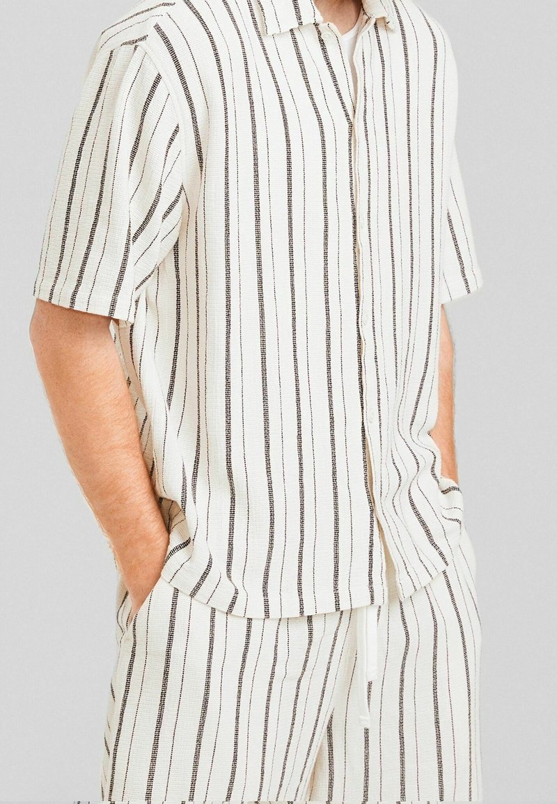 Man som bär en vit kortärmad skjorta med knappar och matchande shorts med tunna vertikala svarta ränder, med händerna i fickorna.