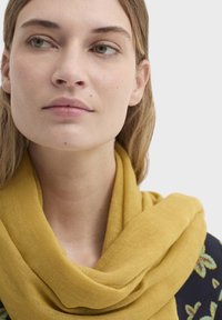 Écharpe en lin jaune enroulée autour du cou, avec une texture douce et un drapé lâche, contrastant avec un haut à motif floral foncé.