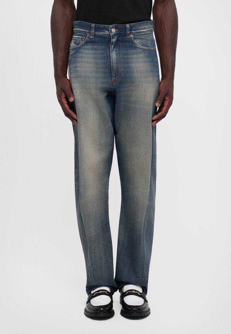 Man som bär blekta rakbenade blå jeans, svarta loafers med guldkedjedetalj och vita strumpor, stående mot en enfärgad bakgrund.