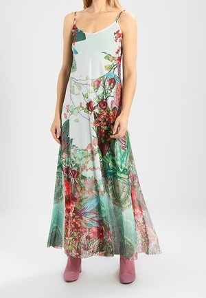 Nainen farkkueväisessä, pitkälti nilkkaan ulottuvassa kukkaisessa mekossa, jossa on vihreitä, punaisia ja sinisiä kasviaiheisia kuvioita, yhdistettynä vaaleanpunaisiin nilkkureihin.