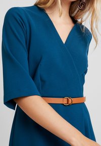 Teal omslagsklänning i texturerad tyg, breda ärmar och brunt läderbälte med en gyllene cirkulär spänne.