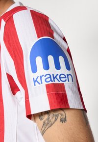 Punainen ja valkoinen raidallinen paita, jossa on sininen Kraken-logo vasemmassa hihassa; sisältää tekstuurista kangasta ja klassisen kauluksen muotoilun.