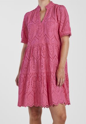 Różowa sukienka z krótkim rękawem, wzorem ażurowej koronki, falowanym dołem i dekoltem w serek, na osobie pokazanej od szyi do kolan.