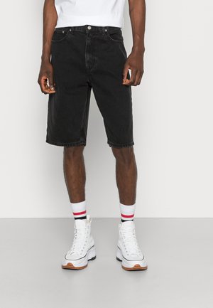 Uomo che indossa pantaloncini di jeans neri al ginocchio, calzini bianchi con strisce rosse e nere e sneakers alte bianche.