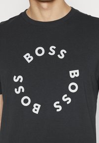 Man som bär en svart T-shirt med vita perforerade bokstäver som stavar "BOSS" arrangerade i ett cirkulärt mönster på framsidan.