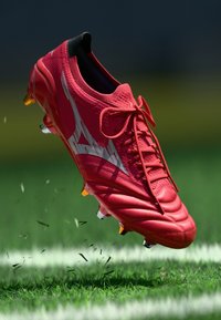 Červené futbalové kopačky s textúrovaným povrchom, šnúrkami a kovovými akcentmi. Obsahujú žlté a biele trny. Okolo je roztrúsené trávnaté znečistenie.