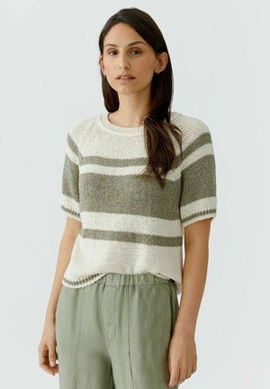 Femme portant un pull en tricot à manches courtes avec des rayures blanches et olives et un pantalon ample vert olive, debout devant un fond uni.