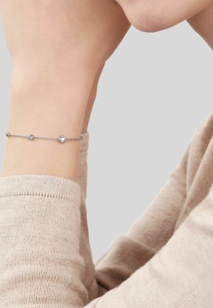 Pulsera delicada de cadena de plata con dos pequeñas piedras preciosas redondas y transparentes, llevada en una muñeca, sobre un fondo neutro.