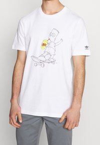 Vit bomullströja med en grafik av en tecknad karaktär på en skateboard som håller en gul dryck. Adidas-logotyp på ärmen.