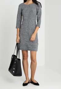 Robe en tricot grise avec un panneau avant texturé, des manches trois-quarts et une coupe ajustée. Associée à un sac fourre-tout noir et des ballerines pointues.