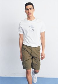 Camiseta blanca con un diseño gráfico y logo en azul, combinada con pantalones cortos cargo en verde oliva. El modelo lleva zapatillas blancas. Fondo simple.