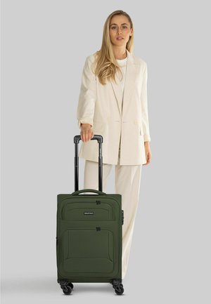 Frau in einem cremefarbenen Anzug, die steht und den Griff eines dunkelgrünen rollenden Koffers auf grauem Hintergrund hält.