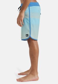 Himmelblaue Boardshorts mit horizontalen Streifen, kontrastierendem blauen Bund und seitlichen Schlitzern für Beweglichkeit. Verfügt über ein schwarzes Logo-Patch.