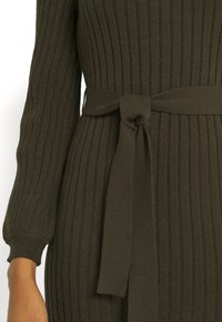 Närbild av en mörkgrön ribbad trikåklänning med lång ärm och knytbart tygbälte i midjan som sitter på en persons överkropp och arm.