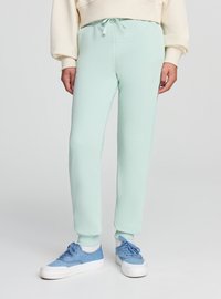 Jasnozielone spodnie dresowe z prążkowanymi ściągaczami przy kostkach, elastyczną talią z sznurkiem, zestawione z niebieskimi trampkami. Miękka tekstura.