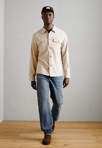 Μπεζ πουκάμισο με κουμπιά και δύο τσέπες στο στήθος, συνδυασμένο με ανοιχτού μπλε τζιν παντελόνι και καφέ παπούτσια. Το μοντέλο στέκεται μπροστά από έναν απλό φόντο.