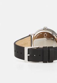 Montre avec bracelet en cuir noir, dotée d'un boîtier en acier inoxydable et de finitions en acier argenté, avec une finition mate lisse et une fermeture à boucle ajustable.