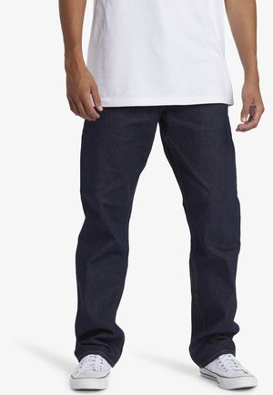Mann trägt dunkelblaue Jeans, weiße Sneakers und ein schlichtes weißes T-Shirt, steht mit entspannten Händen an den Seiten.
