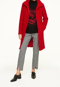 Manteau en laine rouge avec un col structuré, pull noir avec un motif de rose, et pantalon en pied-de-coq gris avec des fentes sur les côtés, accompagné de chaussures noires.