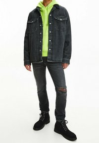 Chaqueta de denim negra con dos bolsillos en el pecho, superpuesta sobre una sudadera verde brillante. Jeans rasgados oscuros y botas negras con cordones completan el conjunto.