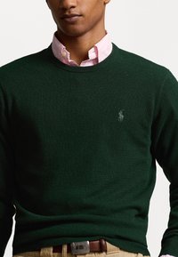 Man draagt een donkergroen trui over een roze overhemd met kraag, beige broek en een bruine leren riem, bijgesneden bij de schouders en taille.