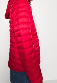 Rote Daunenjacke mit Kapuze, mit gestepptem Design, Reißverschlusstaschen und einer glatten, glänzenden Textur. Über Jeans getragen.