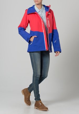 Veste colorée avec des panneaux rouges et bleus, capuche, fermeture éclair devant et deux poches latérales. Portée avec un jean bleu et des bottes beiges.