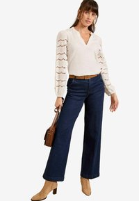 Light beige top with lace detail on sleeves, paired with dark blue wide-leg jeans and tan ankle boots. Brown belt and small brown handbag.