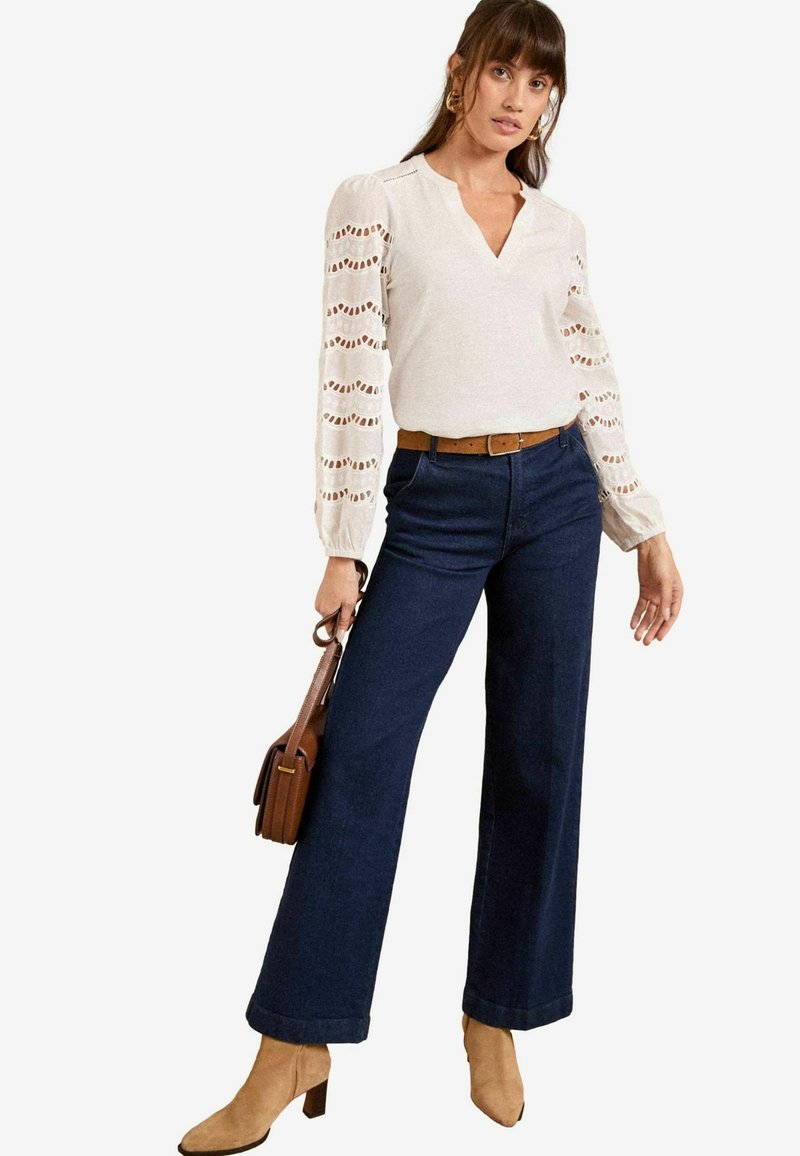 Light beige top with lace detail on sleeves, paired with dark blue wide-leg jeans and tan ankle boots. Brown belt and small brown handbag.