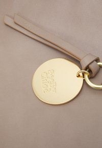 En beige lædertaske med detalje i form af en gylden cirkulær mærkat, hvor "See By Chloé" er præget. Glat overflade med flad rem og metalbeslag.