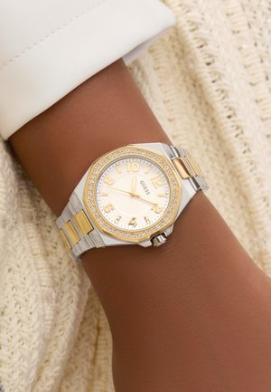 Montre argentée et dorée avec un cadran texturé blanc et des chiffres dorés, entourée d'une lunette sertie de cristaux et d'un bracelet en métal lié.