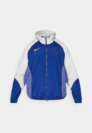 Blauw en witte windjack met een rits aan de voorkant, capuchon en contrasterende paarse accenten; voorzien van een klein oranje logo op de borst.