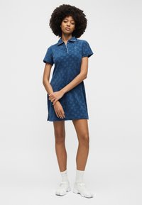 Γυναίκα με σγουρά μαλλιά που φοράει μπλε φόρεμα σε στυλ πόλο, λευκές κάλτσες και χοντρά λευκά αθλητικά παπούτσια, στέκεται μπροστά σε απλό φόντο.