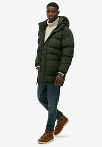 Oliwkowa kurtka puchowa z kapturem, zapięcie na guziki i kieszeniami bocznymi, noszona na jasnym, strukturalnym swetrze, w połączeniu z niebieskimi dżinsami i brązowymi butami.