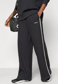 Tamnosive trenerke s bijelim bočnim prugama, izvezenim natpisom "adidas" na džepu, uparen s crnom torbom preko ramena i crnim tenisicama.