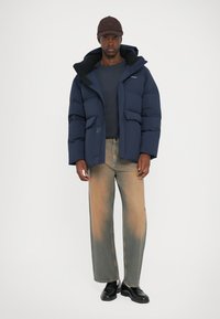 Marineblauwe pufferjack met een ritssluiting aan de voorkant, grote zakken en een capuchon, gecombineerd met licht vervaagde jeans en zwarte loafers, op een neutrale achtergrond.
